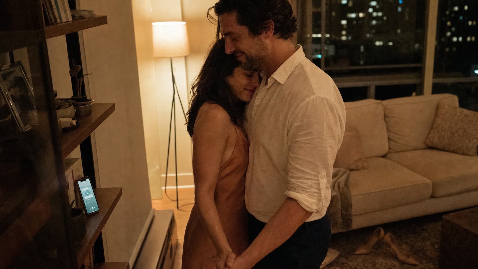 A couple slow dancing in their living room to a song playing from a phone on the shelf